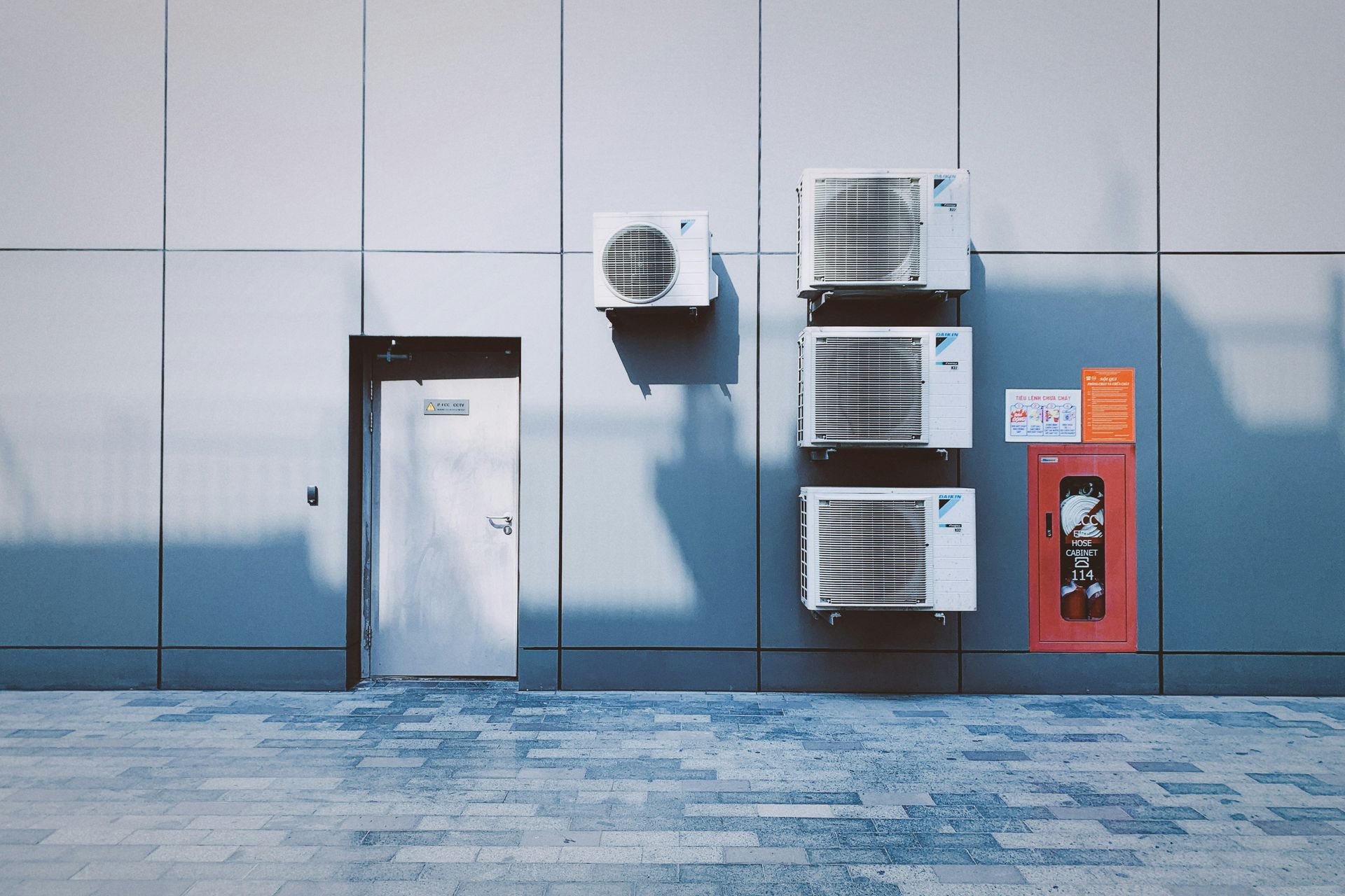 Four air conditioning units on a grey panelled wall beside a red fire cabinet